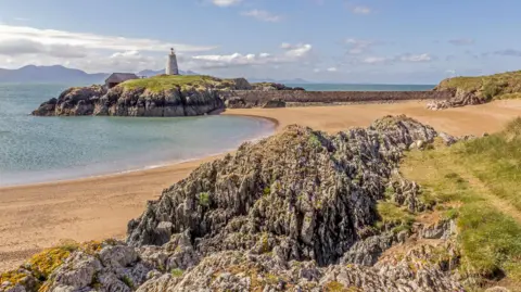 Image shows rocky terrain and a sandy beach in the foreground.
A small island in the background, topped with a lighthouse and an adjacent building.
The island is surrounded by the sea, with mountains visible in the distance.
The sky is mix of blue sky and some clouds above the mountain's on the horizon.