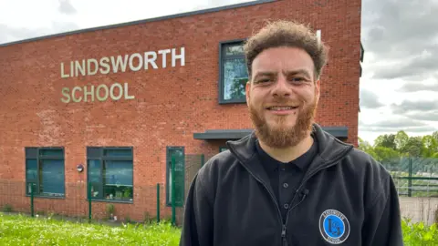 Assistant Headteacher Courtney Ballard stands in front of Lindsworth School, a modern red brick building. He is wearing a fleece with the school logo on it.