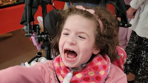 Brown haired 9 year old Thea is sitting on her wheelchair. Ventilator and feeding tube coming out of her neck. Wearing a light pink jumper and a pink neckerchief with red and yellow hearts and blue spots.