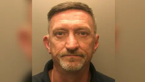 A mugshot of an elder man, looking towards the camera lens. He has grey hair, moustache and a beard. He is wearing a navy blue polo shirt. Stood against a white backdrop.