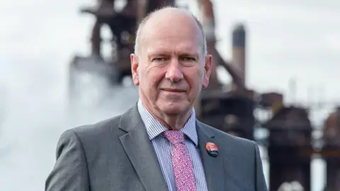 David Rees stood in front of the Port Talbot steelworks with a GMB union badge. He is wearing a suit and a red and white tie.