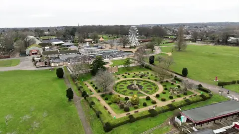 Stephen Huntley/BBC Drone shot of Wicksteed Park showing formal garden laid out as a central circle surrounded by squares with bushes laid in rows. A large pavilion building, other smaller buildings and a big wheel are visible in the background.