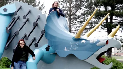 BBC Susie Glatt standing in front of a blue Triceratops that Jude Keenan has climbed