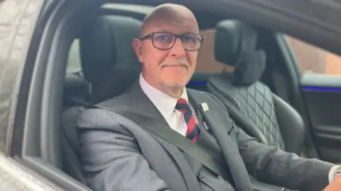 A man in glasses, wearing a three-piece suit and tie, is sat at the wheel of a car. He is looking to the camera and smiling.