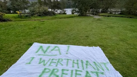 Llinos Alun Protest banner on the grass in front of a lake with the words "Na! i werthiant preifat" in Welsh, meaning "no to a private sale"