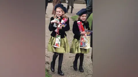 Getty Images Young girls wearing matching outfits of beret-style hats, gold skirts and velvet jackets. They are walking outside.
