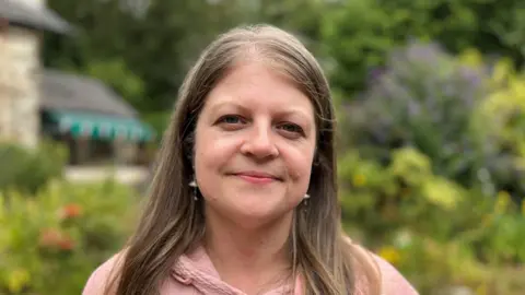 Evie Roberts looking at the camera. She is stood outside the Pensychnant Nature Reserve building and is wearing a pink hooded sweatshirt and has long dark hair. 
