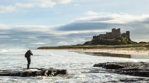 Marcus Hawkins/Getty Images Photographer standing in the surf taking pictures of a cloud-covered Bamburgh Castle on the Northumberland coast 