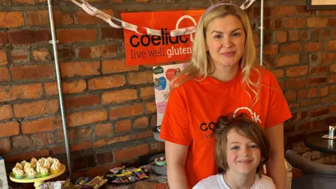 Submit A woman with blonde hair stands with a small boy with brown hair. A bright orange banner hangs behind them that states 'Coeliac UK'. The woman is wearing a bright orange top that matches the banner.