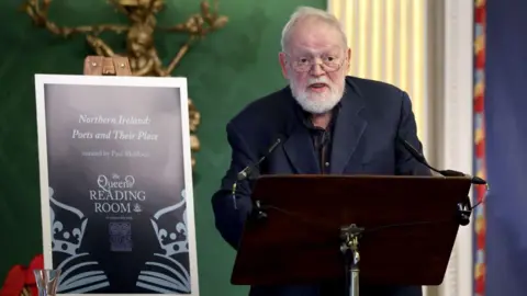 Getty Images Michael Longley, with short white hair and beard, wearing glasses, wears a dark suit and stands behind a lectern. To his right is a sign saying "Northern Ireland Poets and Their Place".