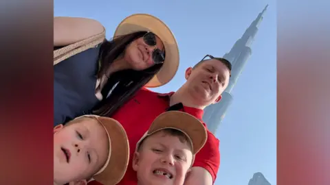 Pete Williams is smiling as he takes a selfie with his partner and two young boys in front of a skyscraper. He is wearing red t-shirt and has short brown hair and a pair of sunglasses over his head. His partner has long straight dark hair and a beige brimmed hat. She is wearing sunglasses and and blue vest. The boys have short hair and are wearing light brown caps. 