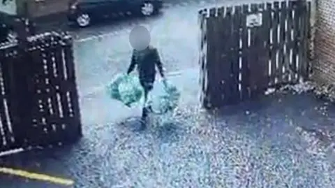 A man carrying four bags of rubbish, two in each hand. He is walking through an open gate into a driveway, with wooden fencing either side. His face is obscured. 