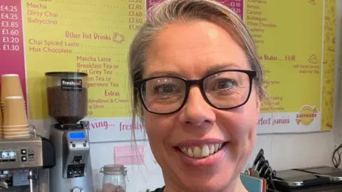 Supplied A selfie-style image of Hannah Barton. She is wearing glasses and smiling while looking into the camera. Behind her is a large yellow menu on the wall and a coffee machine. 