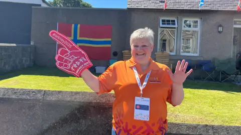 Woman wearing orange top, standing with a red foam hand and smiling