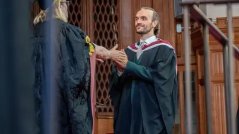 Paul Edwards is wearing academic robes, clasping the hands of the University Vice Chancellor to his left. Mr Edwards has a tie and shirt on under his graduation gown. He has brown hair tied in a ponytail and is smiling. They are in a grand university hall with wooden panelling.