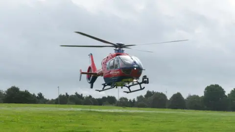 Devon Air Ambulance A red helicopter hovering above a green field on a cloudy day.