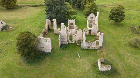 Historic England Archive Aerial view of the ruined Elizabethan house at Hampton Gay. Just a few of the stone walls remain, surrounded by grass and trees.