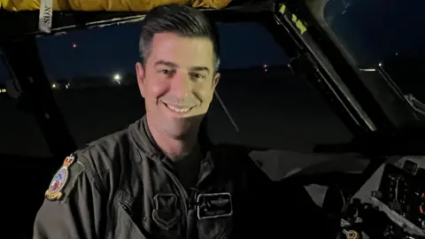 Lieutenant Colonel Michael Devita smiles at the camera in low light as he sits in the cockpit of a bomber plane. He is wearing a khaki or grey bomber jacket with badges on it. His hair is slightly grey.