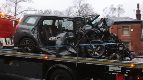A dark Seat car with its front bonnet completely smashed and both right doors torn off is towed away on the back of a lorry past houses.