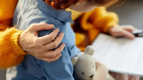 Getty Images Child in a blue shirt, holding a teddy bear with a woman in an orange cardigan putting one arm around the child
