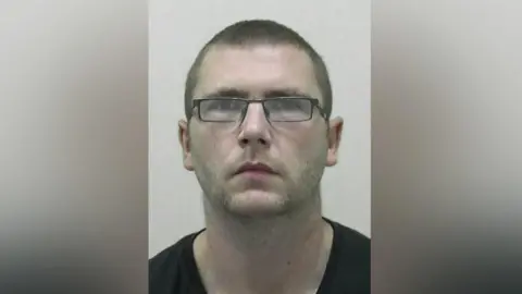 Mugshot of Ian Campbell. he has short dark hair, faint stubble and is wearing a black tshirt and black rectangular framed glasses