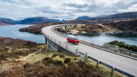 A red and white campervan crosses the Kylesku Bridge. The distinctive curving bridge spans a deep sea inlet.