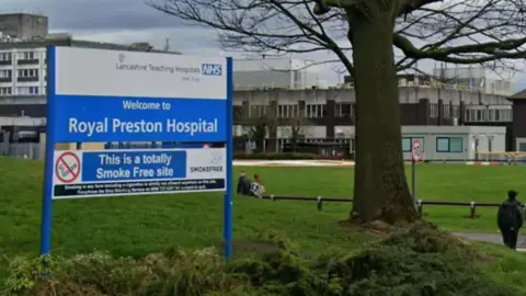 Google The entrance to Royal Preston Hospital showing the welcome sign on a cloudy day. People can be seen walking on the path to the main hospital building.