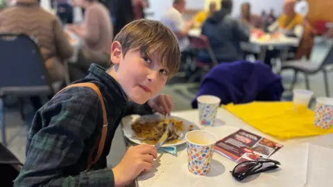 Jon Wright/BBC A 10-year-old boy sits at a table happily eating a plate of food.