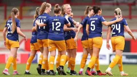 Getty Images Hull City Ladies Football Club wearing navy and amber kits, huddling together on the pitch. Their backs are to the camera and there are names and numbers on the back of their shirts. A red and white viewing area can be seen in the stadium in the background.