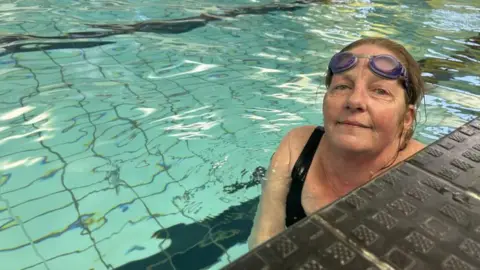 Eleanor Maslin/BBC A woman with brown hair tied back, purple goggles on her forehead and a black swimming costume. She is in a pool and smiling into the camera from the edge. You can see part of the surface of the swimming pool in the background.