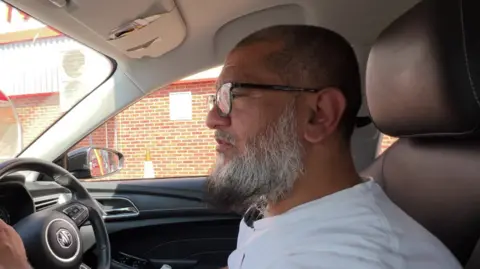 Yasser Araraf sat in his MG car - visible from the steering wheel. He has a mostly white beard and is wearing glasses.