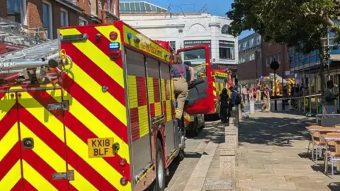 Essex County Fire and Rescue Service View behind fire engines stood in the middle of a road which is on the left. A woman is climbing into the fire engine. In the distance, firemen are standing in the middle of a road which is cordoned off with tape.