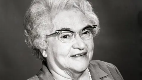 A black and white picture of Gwen Berryman, who is wearing glasses and looking at the camera. She has white hair 