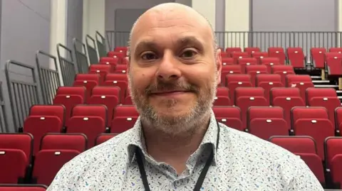 Maddy Jennings/BBC Lee Johnson is looking into the camera, smiling. He has a shirt, grey beard. He is wearing a white and blue patterned shirt and a black lanyard round his neck. Behind him, there are red pull down theatre seats.