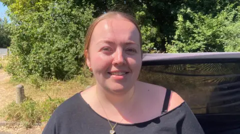 Jess Bayliss has tied back brown hair and is wearing a black top with a gold necklace. 