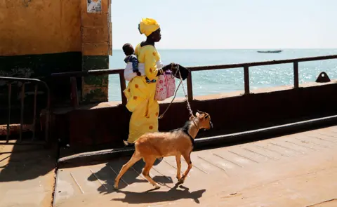 Reuters A woman in bright yellow, carrying a baby and pulling her goat, boards a ferry in Barra, The Gambia - Friday 25 January 2019