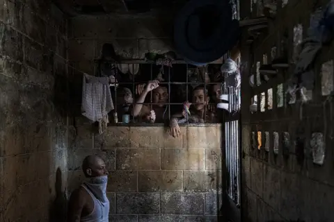 Tariq Zaidi Inmates look out of an overcrowded cell, while a man wears a bandana across his face because of the putrid smell. Penal Center of Quezaltepeque, El Salvador. November 9, 2018.