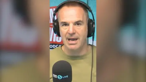 Martin Lewis wearing a green tshirt with a black 5 Live branded mic below him.
