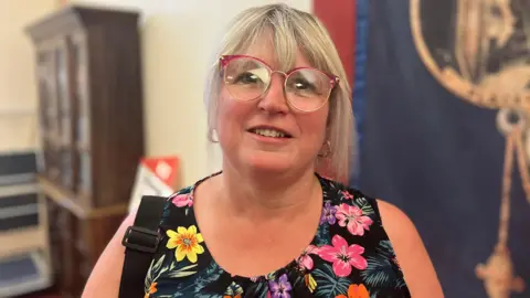 A woman with blonde/grey hair, glasses and wearing a black dress with colourful flowers on it, smiles as she stands in a room with some furniture behind her. She has a black bag strap on her shoulder.
