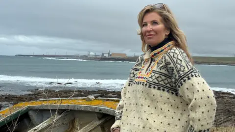 heather jardine wearing a patterned wool sweater standing near a rocky shoreline with waves breaking behind her, an old wooden boat in the foreground, and industrial buildings visible across the water under a cloudy sky.