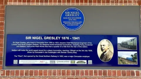 City of Doncaster Council A blue plaque dedicated to Sir Nigel Gresley appears on a wall, with an information board underneath chronicling his history as a railway engineer.