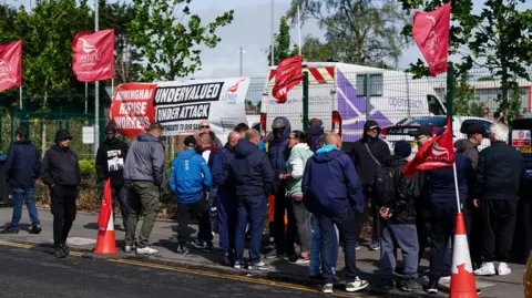 PA Media Workers are on strike outside Perry Barr depot in Birmingham. They are gathered on a pavement, with the area marked with traffic cones. There are red Unite flags and there is a banner that says "undervalued, under attack". Vehicles are parked behind a fence. There are double yellow lines in the road.