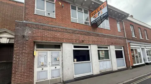 Charlie Jones/BBC The derelict former Barclays bank, which has a brick facade with faded white columns. The door and windows look aged and are slightly cracked. There is an orange, white and black to let sign on the building.