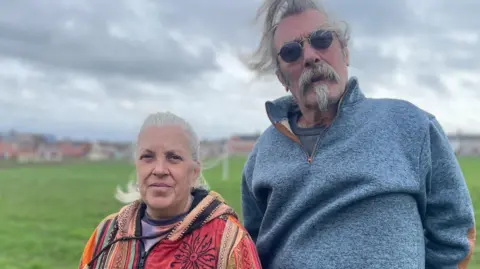 A middle aged couple stand in a large grass field. The woman on the left wears a brightly coloured orange and dark red jacket. The man on the right wears a blue jumper and round tinted glasses.