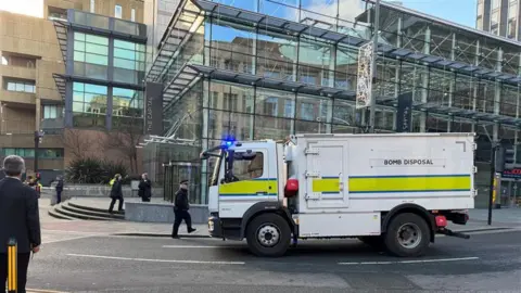 A bomb disposal van parked outside a glass building. Three police officers can be seen outside the building.