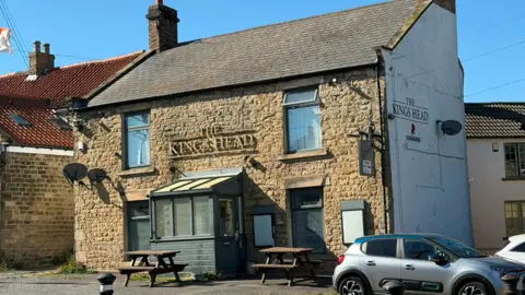 A honey-coloured stone rural pub is illuminated by sunlight against a blue summer sky. The gold signage reads 'The Kings Head'. There are picnic tables and cars parked in front of the building. 