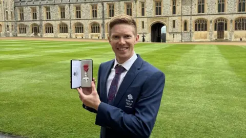 Tom Bosworth A blond-haired man is photographed wearing a blue blazer with a Team GB logo on it. He is looking at the camera, smiling. He is holding up his MBE medal, with a red ribbon, in a black case.