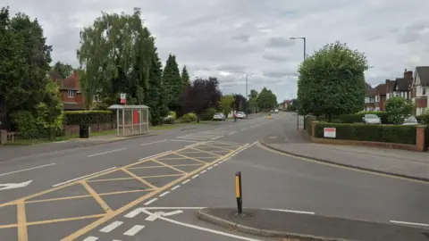 Warwick Road in Solihull. It is a wide road with two lanes and a central reservation. There are residential homes either side of it. Some cars are parked on grassy areas and there is a bus stop too. 