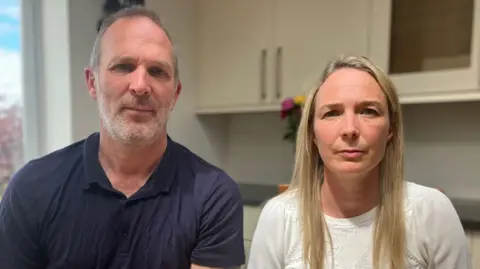 Dr Jack Hawkins sat in the kitchen at home with Sarah Hawkins . He is wearing a dark blue shirt and Sarah has a white top on
