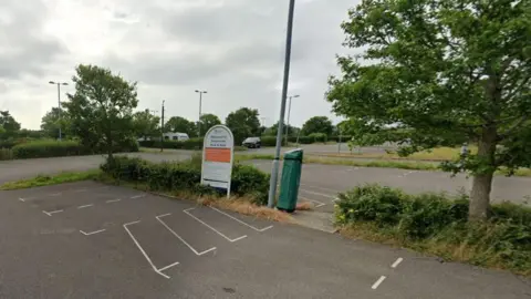 Large car park with vehicles and caravan parked up a sign stands on a verge with low hedging and trees.